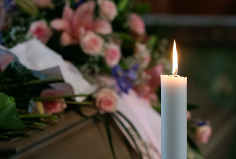 Ett ensamt stearinljus står och brinner framför en kista med blommor
