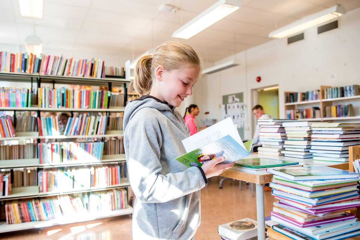 Ett skolbarn står och läser ur en bok i skolbiblioteket och ler.