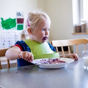 Liten flicka med grön haklapp äter blåbärspaj vid matbordet.