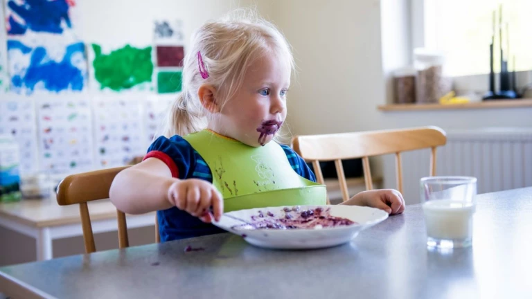 Liten flicka med grön haklapp äter blåbärspaj vid matbordet.