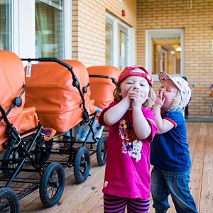 Två barn håller för munnen bredvid en rad orange barnvagnar.