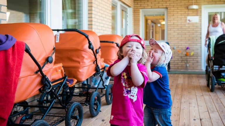 Två barn håller för munnen bredvid en rad orange barnvagnar.