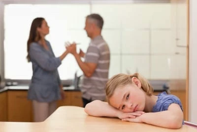 Två föräldrar som står och bråkar och en liten flicka som sitter vid ett köksbord och ser ledsen ut