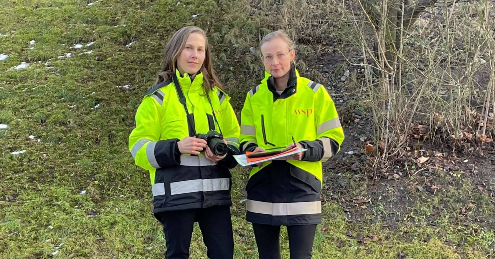 Två kvinnor utomhus ikädda arbetsvästar. Den ena håller i en kamera.