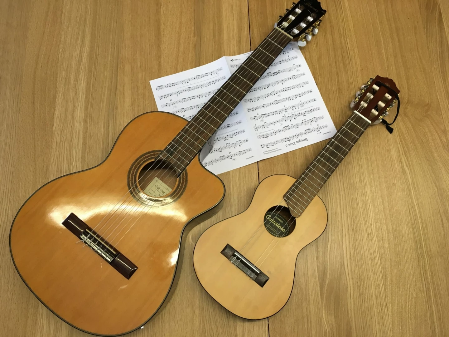 Två gitarrer ligger på golvet tillsammans med ett uppslag notblad. Den ena gitarren är stor och den andra är liten.