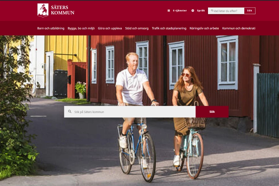 Två personer cyklar på en gata omgivna av röda hus