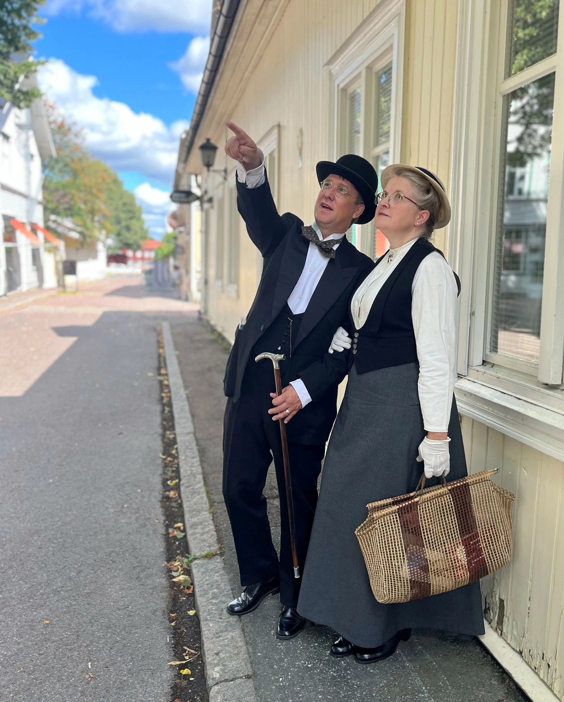 Bilden föreställer två personer, en kvinna och en man. De är klädda traditionsenliga kläder från början av1900- talet. De står på en gata i Säters trästad, mannen pekar på något och kvinnan följer med blicken åt den riktning han pekar i.
