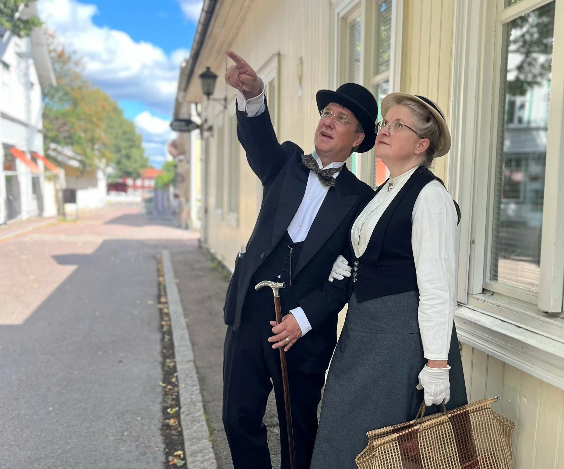 Bilden föreställer två personer, en kvinna och en man. De är klädda traditionsenliga kläder från början av1900- talet. De står på en gata i Säters trästad, mannen pekar på något och kvinnan följer med blicken åt den riktning han pekar i.