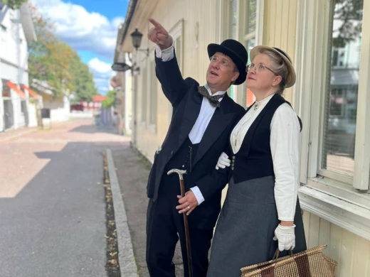 Bilden föreställer två personer, en kvinna och en man. De är klädda traditionsenliga kläder från början av1900- talet. De står på en gata i Säters trästad, mannen pekar på något och kvinnan följer med blicken åt den riktning han pekar i.
