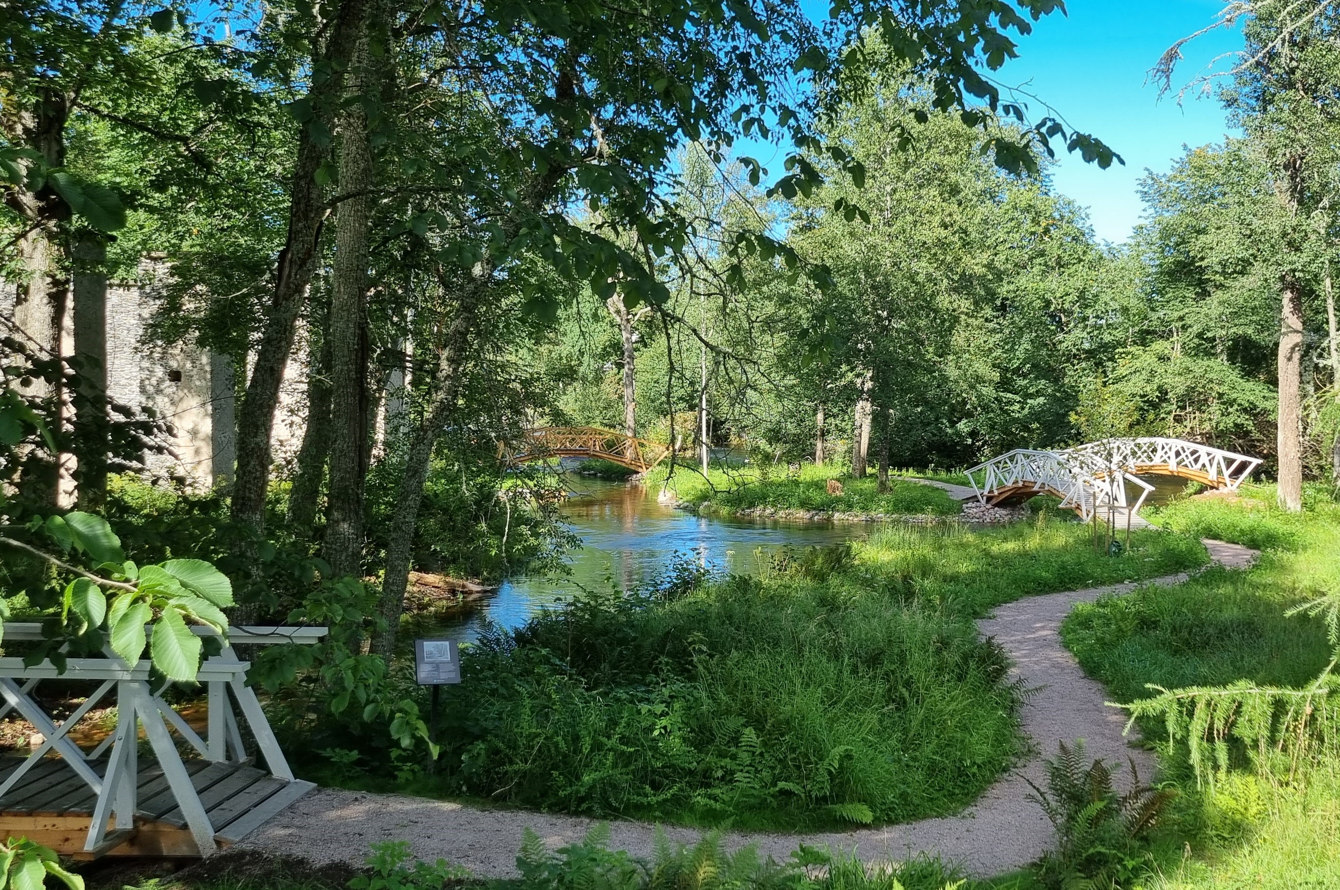Fotot föreställer Engelska parken i Grängshammar. Det är full sommargrönska, solstrålar strilar ner mellan träden. Små vita välvda broar sträcker sig över det porlande vattnet.