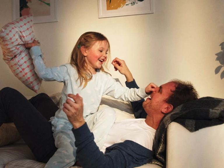 En man ligger på rygg i soffan och ett barn sitter på mannens mage och har kuddkrig.