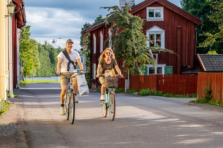 Två personer som cyklar förbi ett fint gammalt hus med en sjö i bakgrunden
