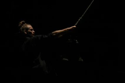 Bild från dansföreställningen Knoster. En man håller i en stor pendel som hänger från taket. scenrummet är mycket mörkt och mannen är svagt upplyst av scenbelysningen.
