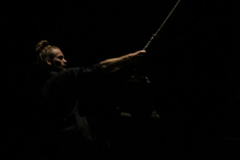 Bild från dansföreställningen Knoster. En man håller i en stor pendel som hänger från taket. scenrummet är mycket mörkt och mannen är svagt upplyst av scenbelysningen.