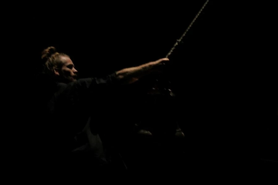 Bild från dansföreställningen Knoster. En man håller i en stor pendel som hänger från taket. scenrummet är mycket mörkt och mannen är svagt upplyst av scenbelysningen.