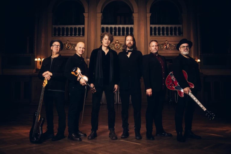 Bilden är en bild på alla medlemar i bandet Engmans Kapell. De står bredvid varandra med sina musikinstrument i handen och tittar in i kameran.