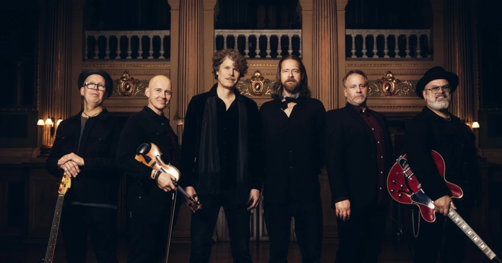 Bilden är en bild på alla medlemmar i bandet Engmans Kapell. De står bredvid varandra med sina musikinstrument i handen och tittar in i kameran.