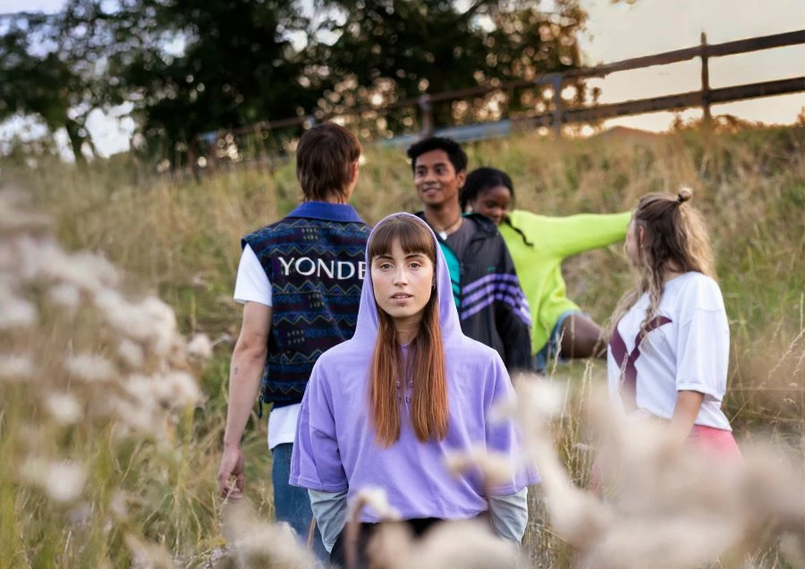 Ett färgfoto på ett gäng ungdomar på en sommaräng. Gräset når dem upp till midjan. En tittar in i kameran. Resten tittar på varandra.