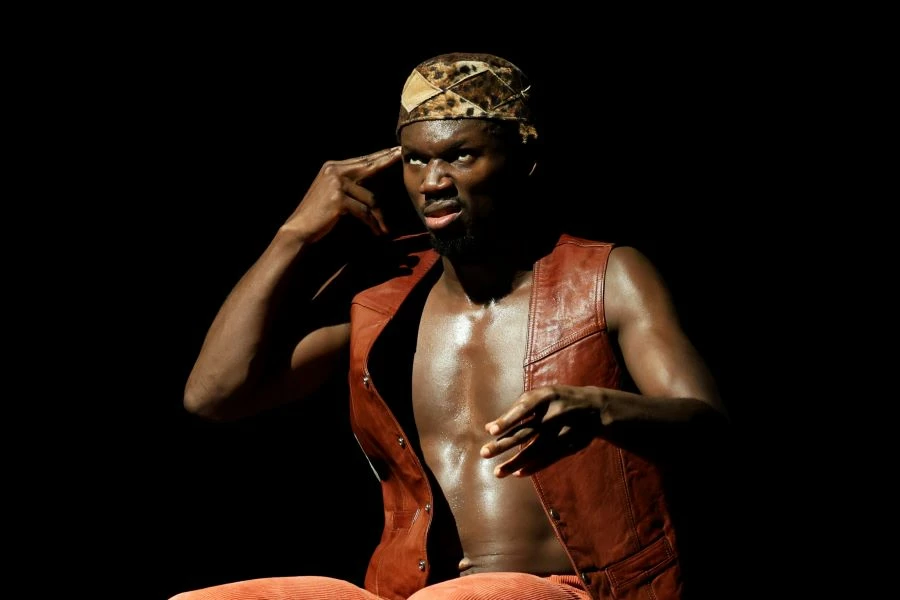 En bild på Dansaren och korografen Ouloy. Han sitter på scenen, allvarlig min, hans egna pekfingrer är riktat mot tinningen.