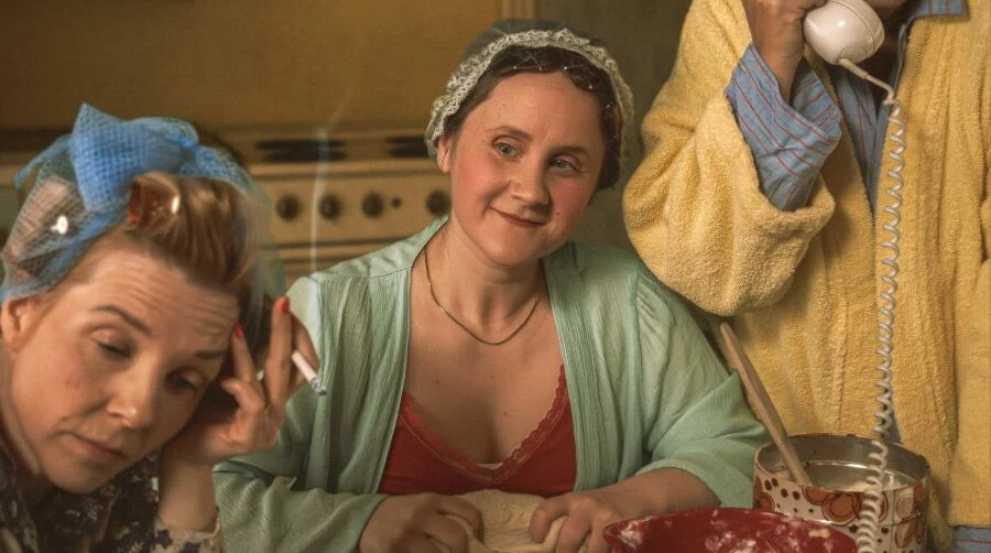 Tre kvinnor vid ett köksbord. Klädda klädda i morgonrockar och papiljotter.