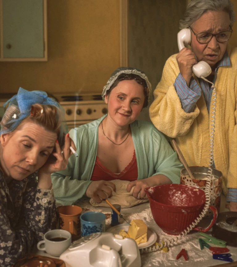 Tre kvinnor vid ett köksbord. Klädda klädda i morgonrockar och papiljotter.