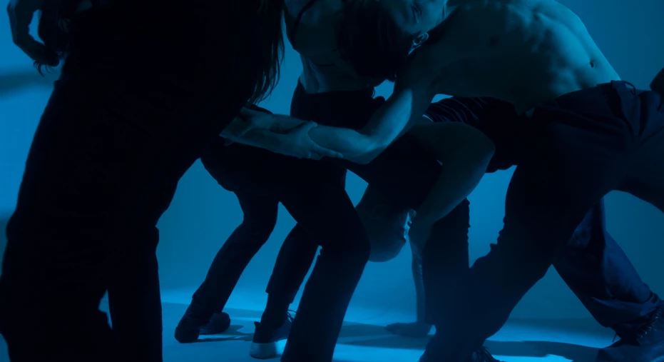 Scenisk bild på kroppar som rör sig på en mörk scen, avtecknas som silutetter mot den ljusa bakgrunden