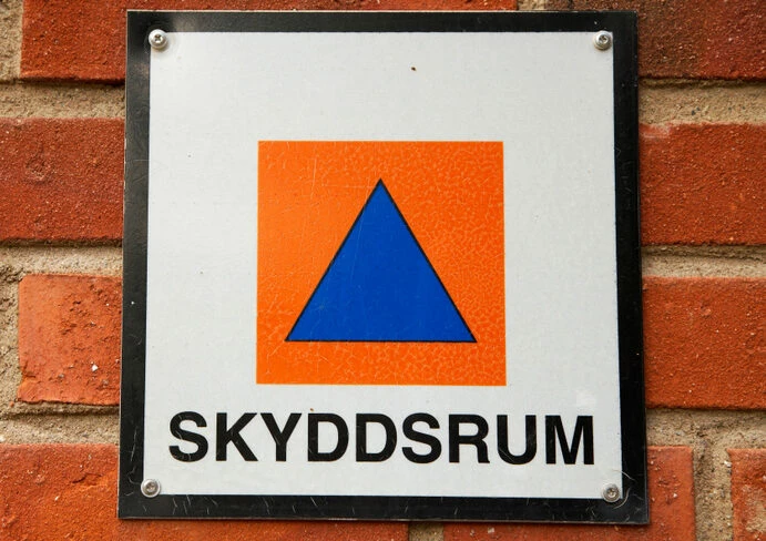 Skylt som visar var ett skyddsrum finns. symbolen är en blå trekant på en orange fyrkant.