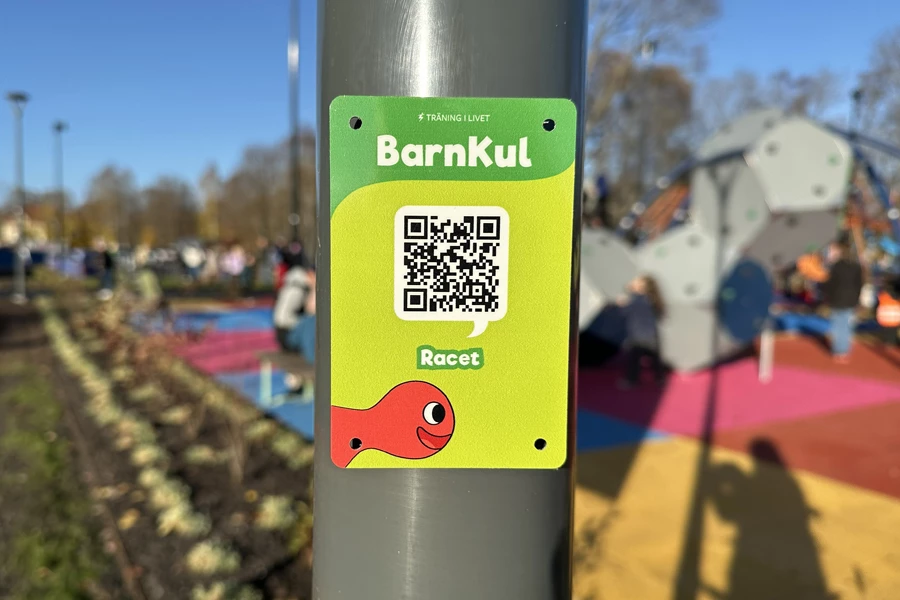 Grön Barnkul-skylt med QR-kod på en stolpe, med parkens färgglada lekytor i bakgrunden.